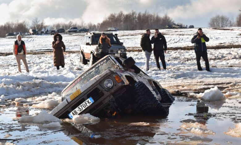 An off-road SUV is stuck in muddy snow with people standing nearby in Gjinari, Albania.