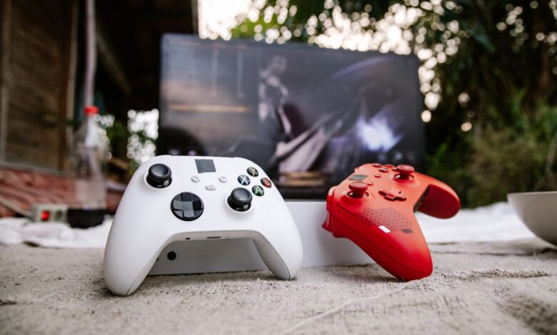 White and red Xbox wireless controllers set outdoors. A computer screen is blurred in the background.