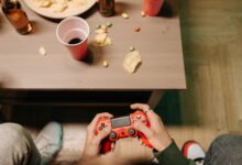 Person holding a red game controller with snacks and drinks around, top view.