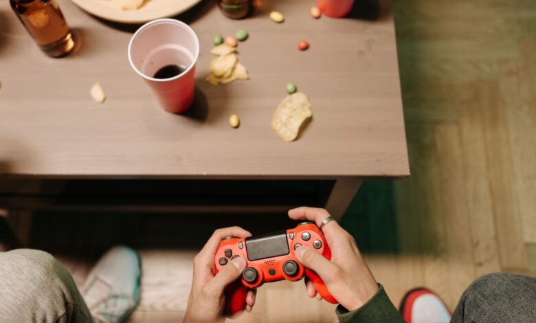 Person holding a red game controller with snacks and drinks around, top view.