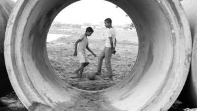 Two boys play soccer through a large cement pipe, showcasing childhood joy in urban settings.