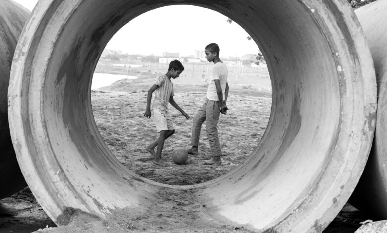 Two boys play soccer through a large cement pipe, showcasing childhood joy in urban settings.