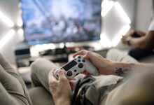 Hands holding a video game controller in a modern living room, playing on TV.