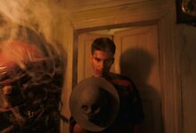 A man in a dimly lit room holds a hat adorned with a skull, surrounded by smoke.