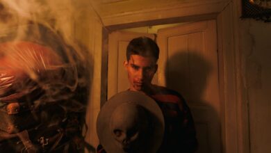 A man in a dimly lit room holds a hat adorned with a skull, surrounded by smoke.