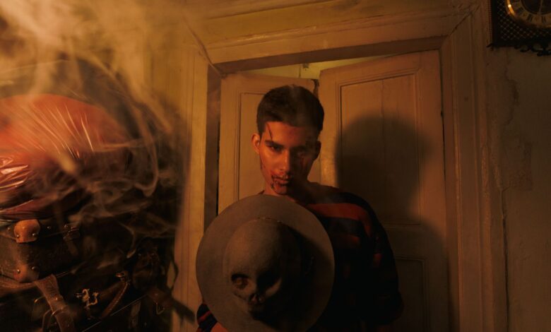 A man in a dimly lit room holds a hat adorned with a skull, surrounded by smoke.