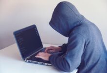 Side view of unrecognizable hacker in hoodie sitting at white table and working remotely on netbook in light room near wall