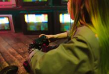 A woman with a prosthetic hand playing video games indoors amidst vintage TVs, showcasing innovation and leisure.