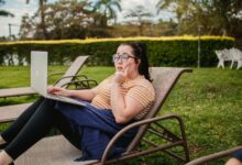 Woman relaxing on lounge chair with a laptop in a lush green garden, enjoying a sunny day.
