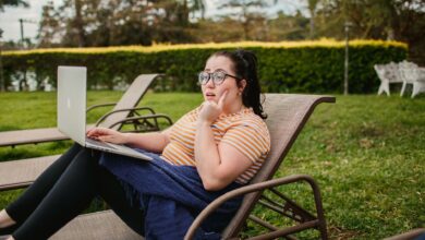 Woman relaxing on lounge chair with a laptop in a lush green garden, enjoying a sunny day.