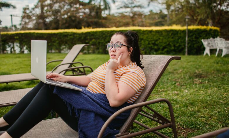 Woman relaxing on lounge chair with a laptop in a lush green garden, enjoying a sunny day.
