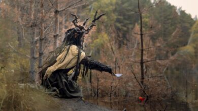 An enigmatic figure with antler headdress sits in a mystical forest clearing.