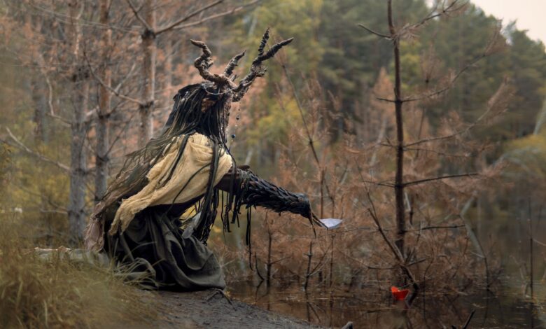 An enigmatic figure with antler headdress sits in a mystical forest clearing.