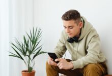 Teenage boy in hoodie reading smartphone with headphones around neck indoors.