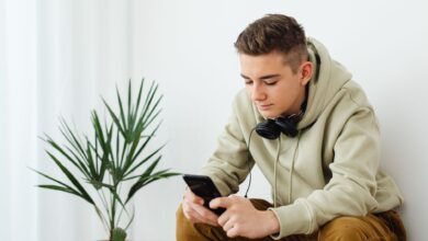 Teenage boy in hoodie reading smartphone with headphones around neck indoors.