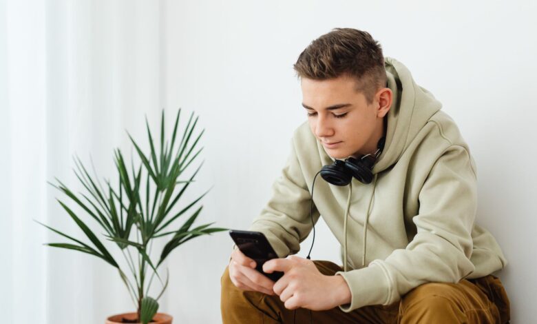 Teenage boy in hoodie reading smartphone with headphones around neck indoors.