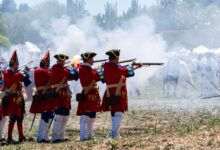 Reenactors in vibrant uniforms firing muskets during a historical battle event.