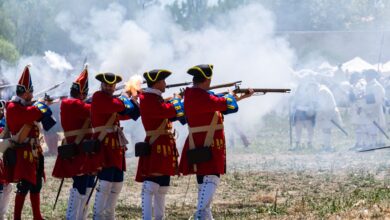 Reenactors in vibrant uniforms firing muskets during a historical battle event.