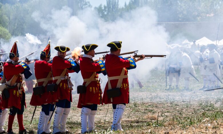 Reenactors in vibrant uniforms firing muskets during a historical battle event.