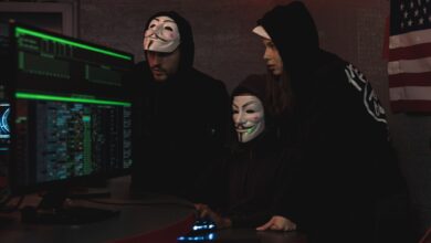 Masked hackers in hoodies working on a system in a dimly lit room, representing cyber security threats.