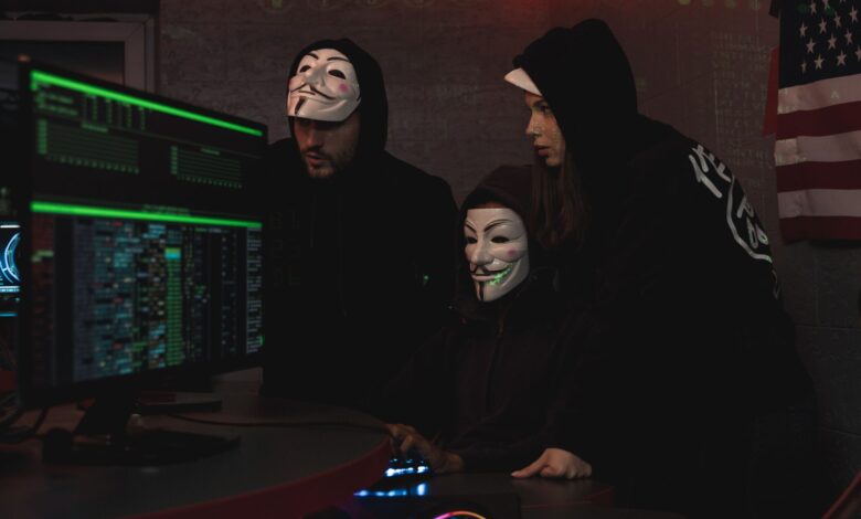 Masked hackers in hoodies working on a system in a dimly lit room, representing cyber security threats.