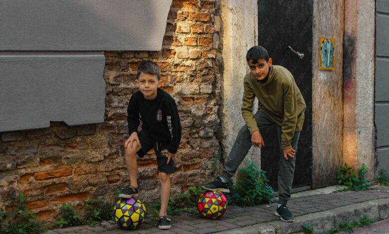 Two boys playing with soccer balls on a rustic urban street, enjoying outdoor fun.
