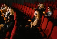 People wearing 3D glasses and eating popcorn in a cinema with red chairs.