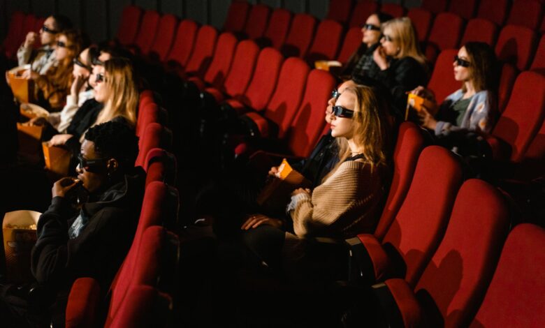 People wearing 3D glasses and eating popcorn in a cinema with red chairs.
