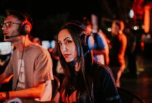 Woman gaming in esports tournament, wearing headset with focused expression, dimmed indoor lighting.