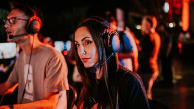 Woman gaming in esports tournament, wearing headset with focused expression, dimmed indoor lighting.