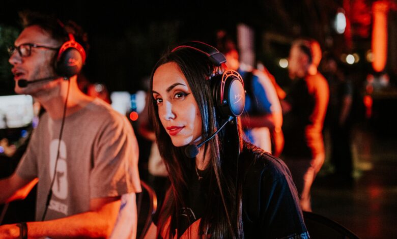 Woman gaming in esports tournament, wearing headset with focused expression, dimmed indoor lighting.