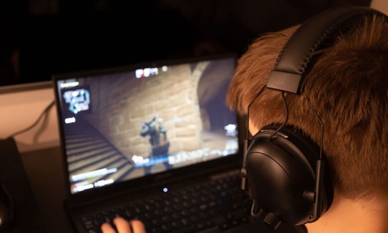 Teenage boy wearing headphones focused on playing a video game on a laptop screen indoors.