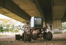 Old damaged vehicle with vintage television set on rough pathway under bridge in sunlight