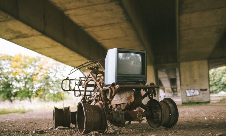Old damaged vehicle with vintage television set on rough pathway under bridge in sunlight