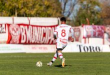 A soccer player in a white uniform dribbles the ball during a sunny outdoor match.