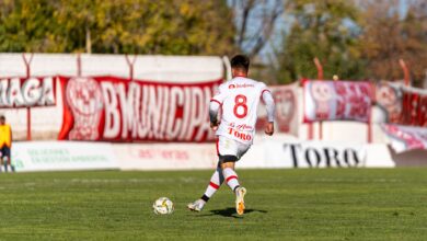 A soccer player in a white uniform dribbles the ball during a sunny outdoor match.