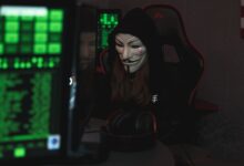 A mysterious hacker wearing a Guy Fawkes mask and black hoodie in a dimly lit room focused on computer screens.