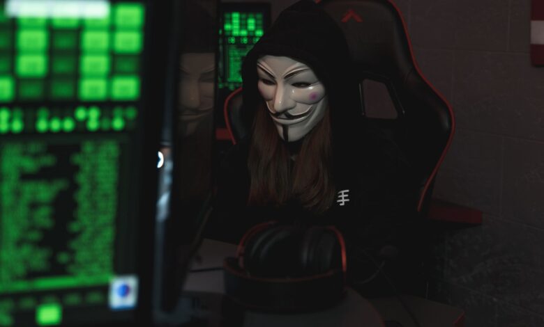 A mysterious hacker wearing a Guy Fawkes mask and black hoodie in a dimly lit room focused on computer screens.