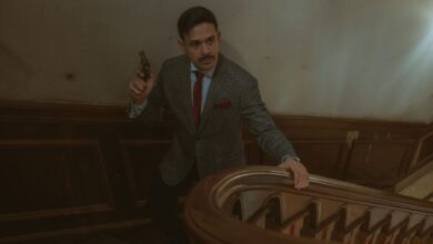 Moody scene of a man in a suit with a revolver on an ornate staircase.
