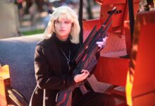 A blonde cosplayer poses with a prop weapon in a vivid outdoor setting, capturing artistic cosplay charm.