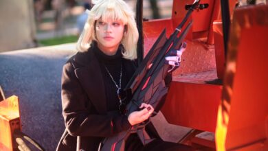 A blonde cosplayer poses with a prop weapon in a vivid outdoor setting, capturing artistic cosplay charm.