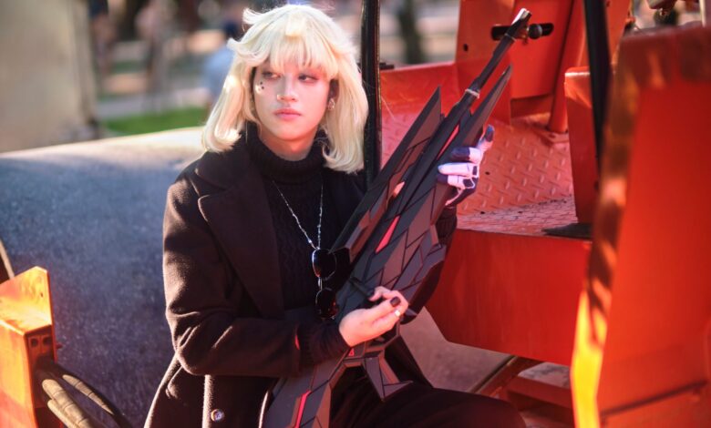 A blonde cosplayer poses with a prop weapon in a vivid outdoor setting, capturing artistic cosplay charm.