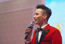 Asian male emcee in a stylish red suit holding a microphone, speaking at an indoor event.