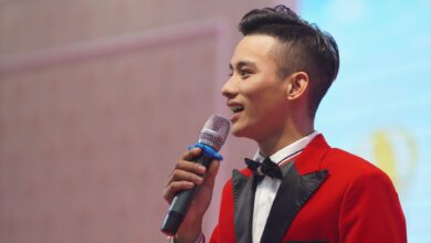 Asian male emcee in a stylish red suit holding a microphone, speaking at an indoor event.