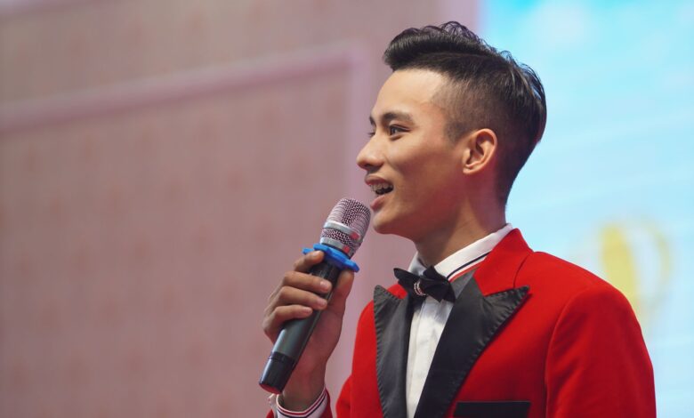 Asian male emcee in a stylish red suit holding a microphone, speaking at an indoor event.