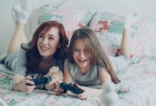 A mother and daughter playing video games on bed, smiling brightly.