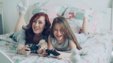 A mother and daughter playing video games on bed, smiling brightly.