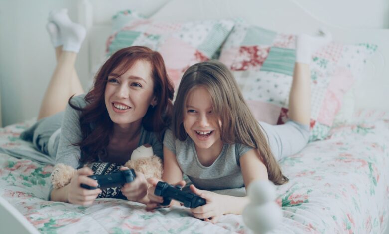 A mother and daughter playing video games on bed, smiling brightly.