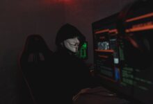 An anonymous hacker wearing a mask working on a computer in a dark room.