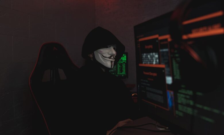 An anonymous hacker wearing a mask working on a computer in a dark room.